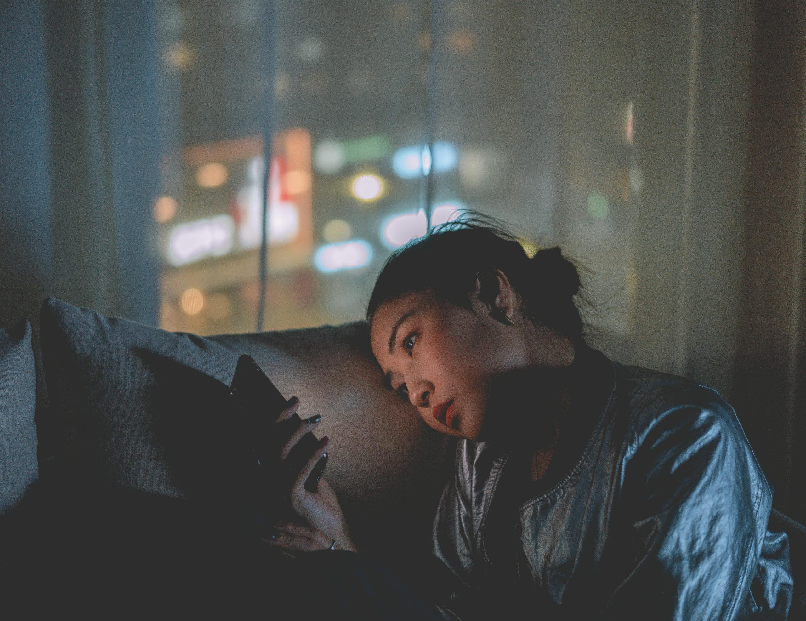 A woman looking at her mobile phone at night with city lights blurred in the background.