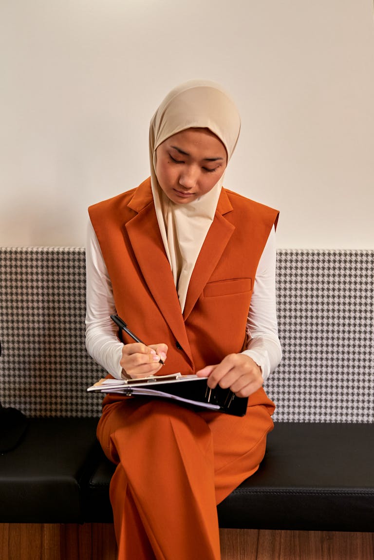 A hijabi woman in an orange suit sits in a modern office taking notes.
