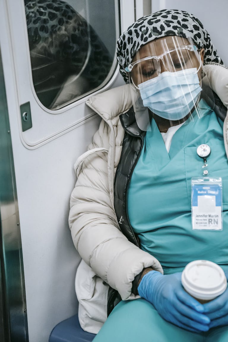 Tired from require overtime African American nurse in warm clothes and protective mask sleeping in metro train with coffee to go in hands