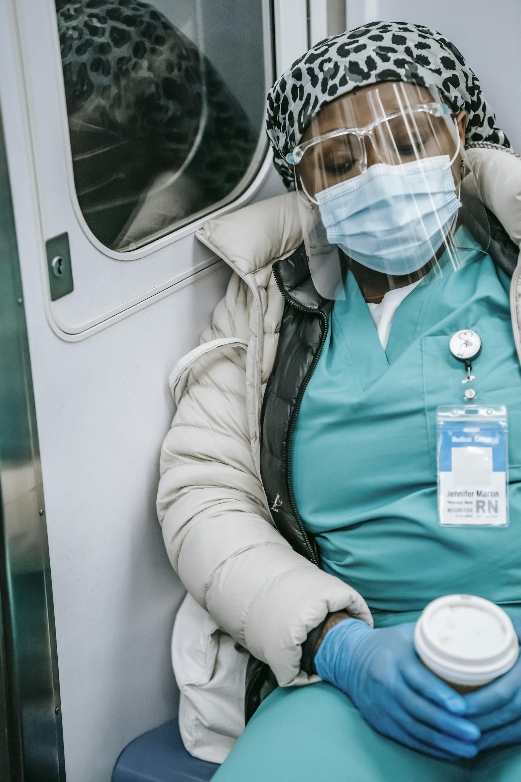 Tired from require overtime African American nurse in warm clothes and protective mask sleeping in metro train with coffee to go in hands