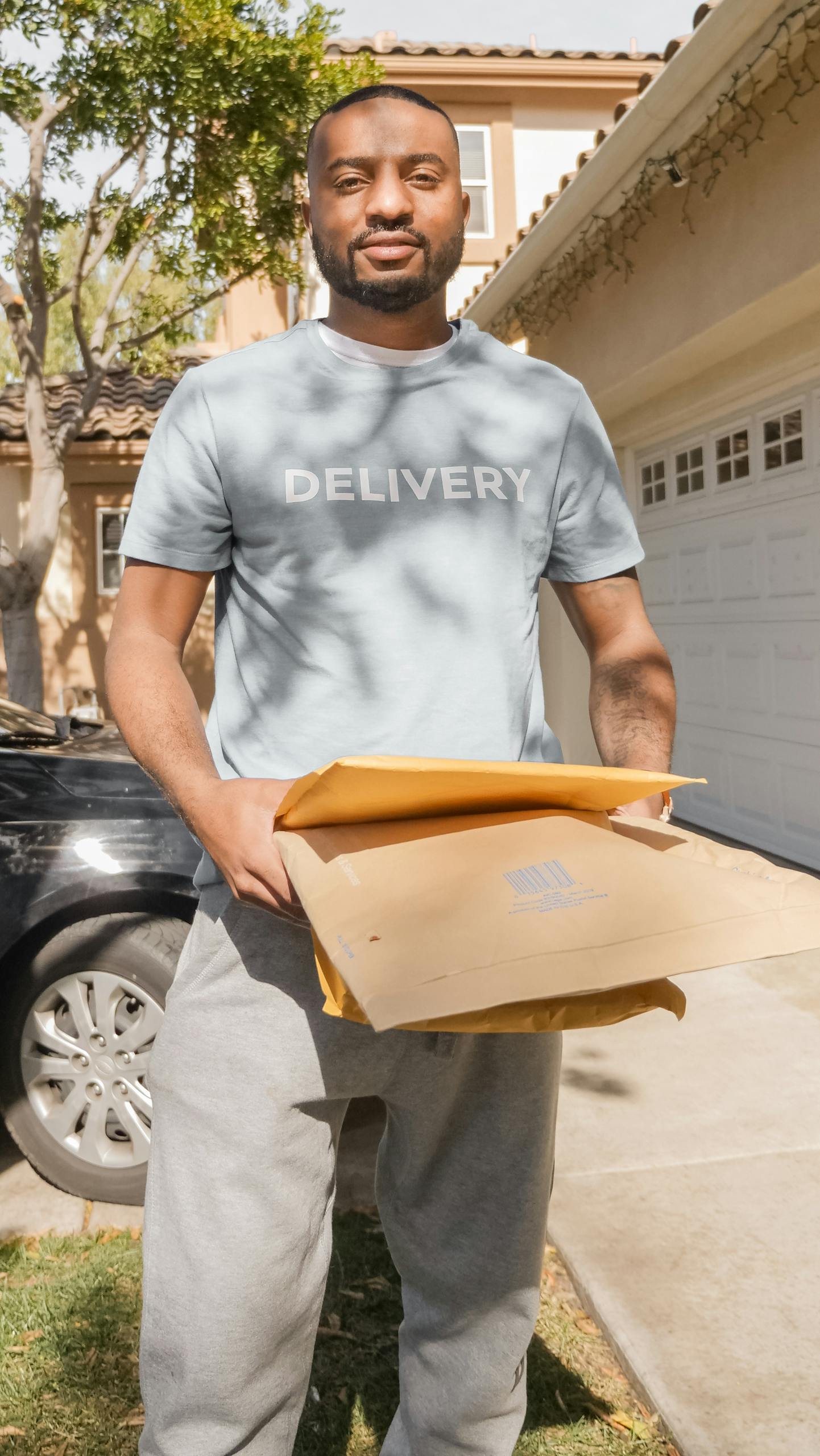 A delivery man holding parcels outdoors on a sunny day near a house and car.