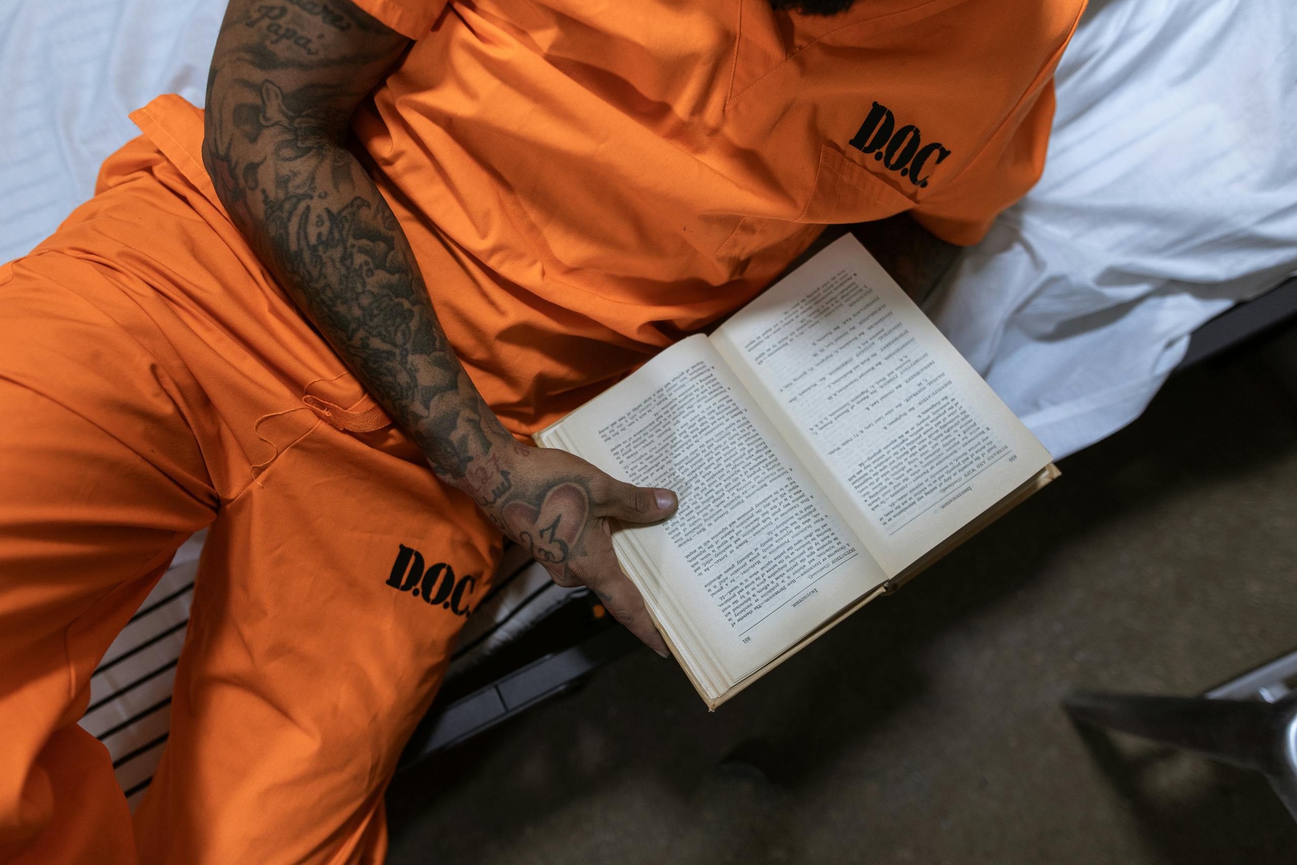 Tattooed inmate reading a book on a bed in a prison cell.