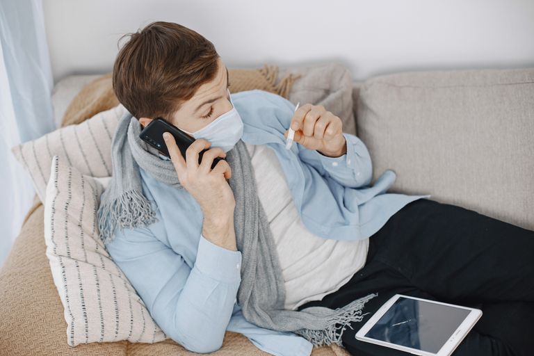 Man using telehealth services while sick at home. Lying on sofa with phone and thermometer.
